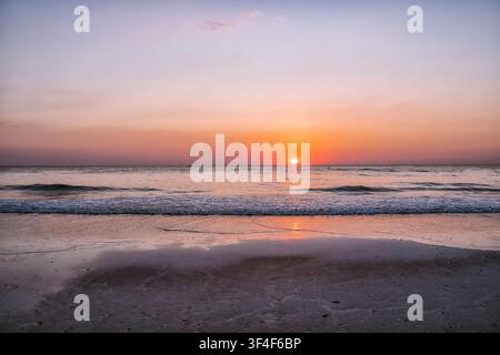 Sun dips low, painting sky in soft orange and pink. Waves gently roll, leaving foam on damp sand. Silent, peaceful beach holds still as day ends. Perf Stock Photo