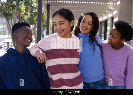Diverse teenage friends chatting and smiling under pergola on campus with knit sweater, navy hoodie Stock Photo