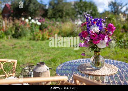 Close up of flower arrangement put on glass table in summer garden. Lounge zone with furniture and bouquet of fresh blooms in vase Stock Photo