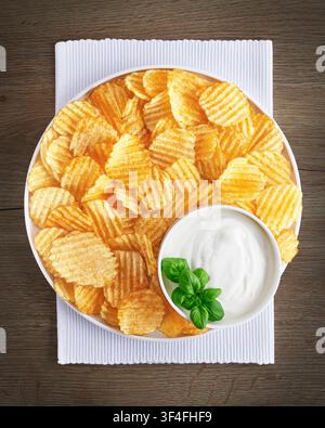 Ruffled potato chips served on a round plate with sour cream dipping sauce garnished with fresh green basil on white napkin over wooden background. Stock Photo