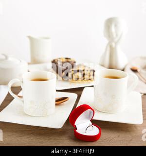White gold diamond engagement ring in red velvet heart box on table with two coffee cups, chocolate cake slices and ceramic figurine. Stock Photo