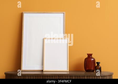 Two blank wooden picture frames and decorative vases on a dark wooden cabinet against an orange wall, a large amber glass vase and a smaller dark glas Stock Photo