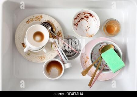 Pile of dirty dishes, cups, bowls, and cutlery in a white kitchen sink with a green sponge, top view Stock Photo
