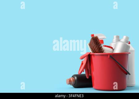 Red bucket filled with cleaning supplies, including spray bottles, brush, and gloves, on a light blue background Stock Photo