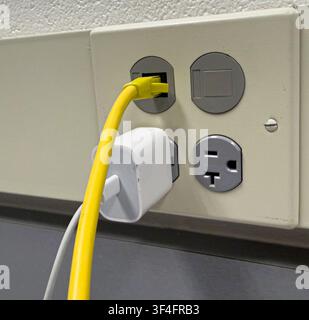 Beige wall features three electrical outlets. Yellow Ethernet cable plugs into one socket. White USB charger connects to another outlet. Gray surface Stock Photo