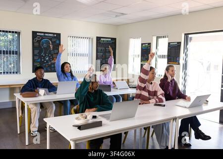Diverse youth classmates raising hands and working on laptops in classroom with headphones Stock Photo