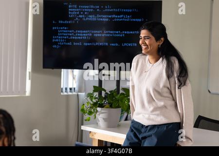 Adult Indian woman in tan top presenting code in office, monitor, plant, whiteboard, copy space Stock Photo