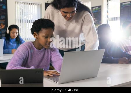 Indian teacher leaning over African teen wearing lavender sweater typing on laptop in classroom Stock Photo