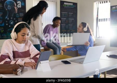 Diverse female students sitting at study table using tablet on white stand wearing headphones Stock Photo