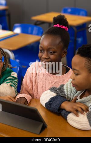 African American kids smiling and viewing tablet at school desk, girl in pink sweater and scrunchie Stock Photo