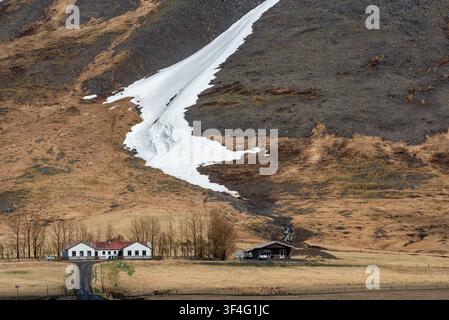 Iceland landscape of a paved country road with a wooden fence winding through a dry, golden grassy field towards a small white, red-roofed farmhouse s Stock Photo