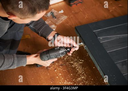 Handyman tightening screw in metal plate on wood surface with drill, close up of detailed work. Indoor workshop with chips and equipment, furniture assembly and fixing concept Stock Photo