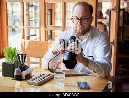 Bearded man adjusting settings on digital camera in cafe, preparing for shooting, creative workflow, professional photography equipment, freelance lifestyle and content production Stock Photo