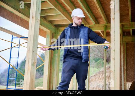 Man using tape measure to assess distance within wooden house skeleton, inspecting spacing and build accuracy during development Stock Photo