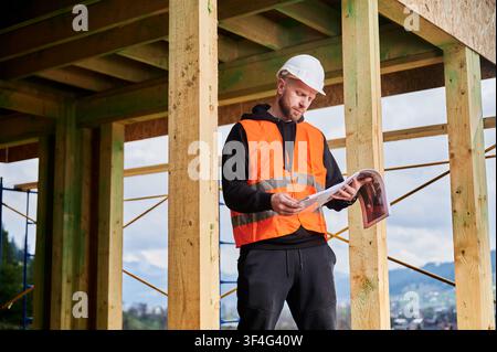 Foreman checks architectural drawings within unfinished wooden house, supervising development and controlling quality during construction process Stock Photo