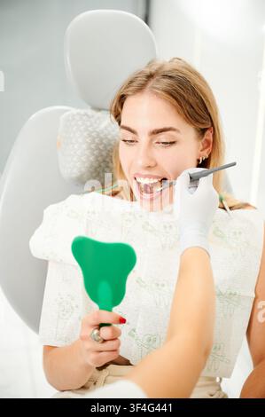 Woman checks teeth in green hand mirror during dental exam. Professional oral inspection in clinic chair with mirror tool. Useful for dentistry advertising, patient care, hygiene promotion Stock Photo