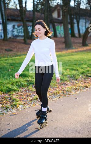 Happy female roller skater performing controlled glide on park path, relaxed expression and athletic pose in sunlight. Suitable for active lifestyle, wellness, energy or hobby use Stock Photo