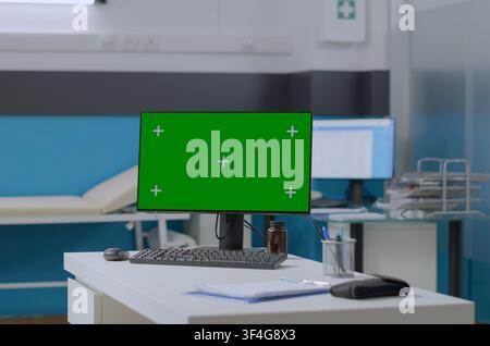 Green screen computer with isolated display stands on a table in an empty hospital office, leaving copy space for a replaceable medical interface. Stock Photo