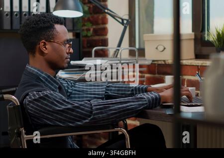 Employee with special needs uses a laptop to analyze ecommerce statistics, creating a financial development project while seated in a wheelchair at work. Stock Photo