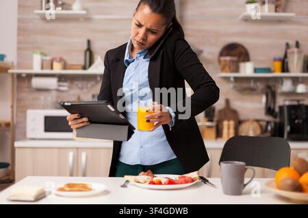 Multitasking woman talks on the phone while holding a tablet and drinking juice, balancing breakfast and work pressure in the kitchen. Stock Photo