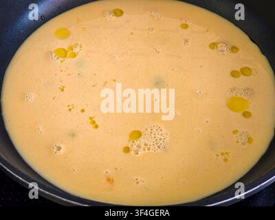 In a kitchen, a non stick pan contains a raw beaten egg mixture. Olive oil droplets float on top while air bubbles form, indicating cooking for breakf Stock Photo