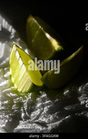 A high-angle macro photograph of fresh green lime wedges placed on a crumpled white paper napkin. Directional lighting emphasizes the translucent, juicy texture of the citrus pulp and highlights the dramatic folds of the napkin. Ideal visual asset for culinary concepts, cocktail recipes, food styling, bar menus, refreshing summer drinks, and healthy nutrition backgrounds. Stock Photo