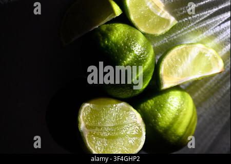 A moody, low-key macro photograph featuring whole green limes alongside fresh halves and wedges on a dark background. Strong directional lighting creates bright highlights on the textured peel and juicy pulp while casting deep, dramatic shadows. Perfect imagery for cocktail bar menus, citrus branding, food photography, tropical drink concepts, and culinary design. Stock Photo
