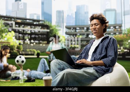 Asian student smiling and writing notes on laptop while relaxing on beanbag in college campus, typing on computer with coffee cup nearby. Independent young man studying alone. Stock Photo
