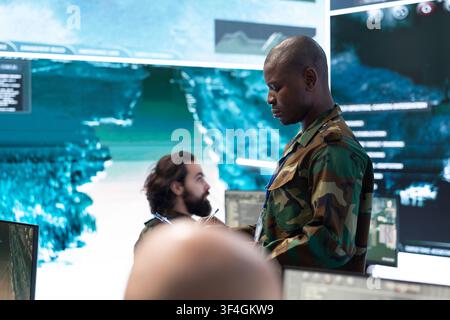 Close up of army officer analyzing operation data with tablet, walking by surveillance screens in command center. Military team leader coordinates enemy target tracking and intelligence updates Stock Photo