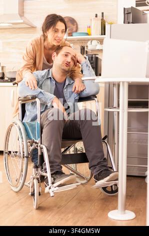 Handicapped man in a wheelchair and his wife take part in a laptop video conference from the kitchen, showing online communication at home, inside an accessible domestic kitchen. Stock Photo