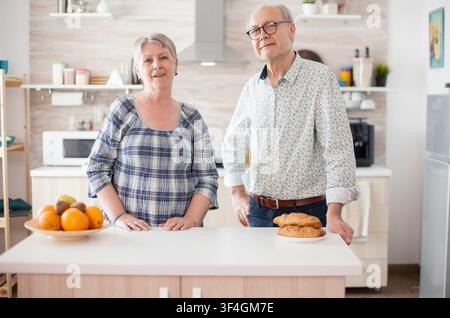 Cheerful older couple looks at the camera in a kitchen while preparing breakfast, showing happy retirement, domestic togetherness and morning routine. Stock Photo