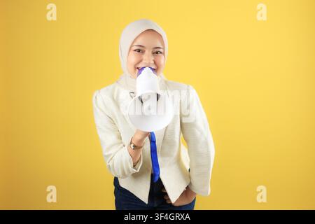 Young Asian Woman in Hijab or Veil Happily Holding Megaphone, Looking at the Camera, Isolated on Yellow Background Stock Photo