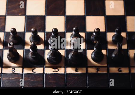 Overhead detailed photograph of black wooden chess pieces and pawns lined up on a dark checkered chessboard with visible algebraic rank and file notations. Dramatic warm lighting creates deep contrast, representing strategic execution, leadership, authority, corporate tactics, and critical decision-making. Stock Photo