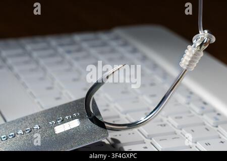 Fishing hook piercing a credit card over a blurred laptop keyboard. Online phishing scams, cybersecurity threats, digital fraud Stock Photo