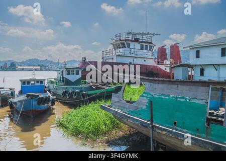 Several weathered wooden motorboats and a large red tugboat are anchored along the grassy bank of the Mahakam River under a bright cloudy sky. Stock Photo