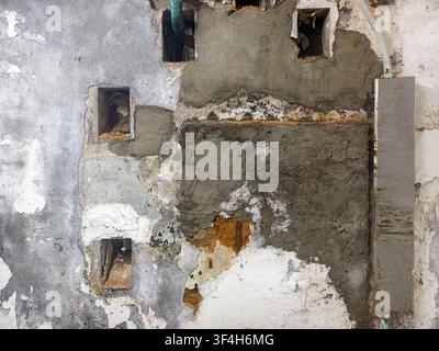 damaged, uneven wall undergoing rough masonry repairs with exposed cavities, layers of old plaster, peeling paint, and fresh cement patches Stock Photo