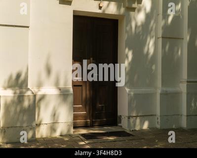Front view of a classic dark brown wooden double door on a light cream building wall with beautiful summer tree leaf shadows. Stock Photo