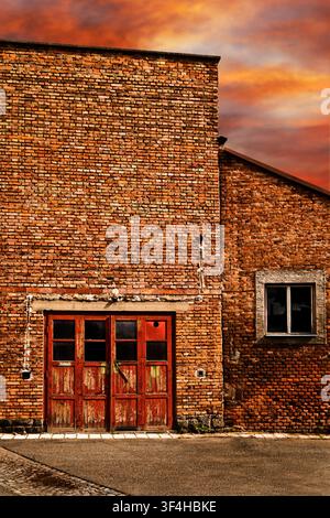 Weathered red brick industrial building with worn doors and windows beneath a dramatic orange sunset sky Stock Photo