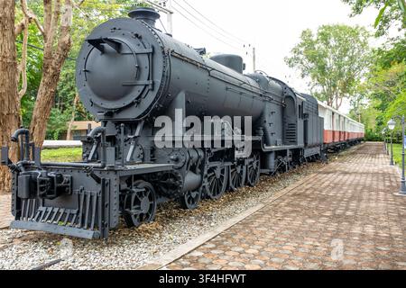 Vintage black steam locomotive with red and white carriages on display track surrounded by trees and paved path Stock Photo