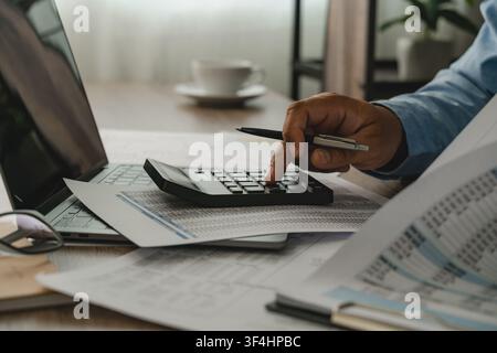 man presses a calculator over accounting reports beside a laptop, reviewing financial figures with a pen on a desk for report preparation, bookkeeping Stock Photo