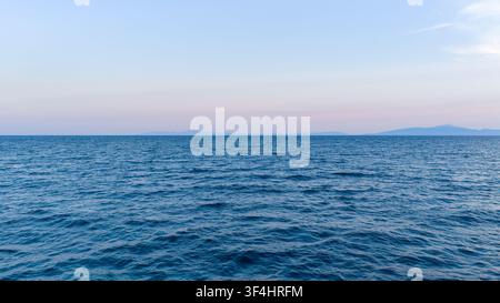 Gentle blue waves stretch toward a low distant coastline beneath a clear pastel sky, creating a tranquil seascape near Nea Moudania, Greece. Stock Photo