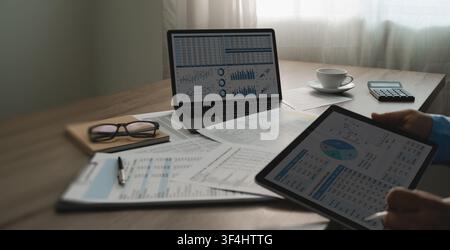 Businessman review annual financial spreadsheets on tablet beside laptop charts, calculator, printed reports, comparing digital data for document chec Stock Photo