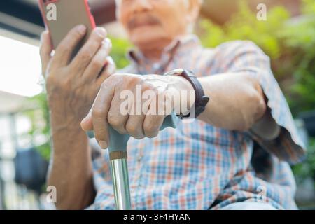 Close up hand of senior man with a beard holding a walking stick while using phone outside in sunlight. Senior health and lifestyle concept. Stock Photo