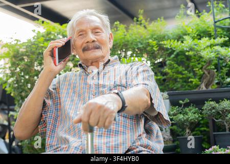 Portrait image of smiling senior man with walking cane and using mobile phone while sitting in the garden at home. Stock Photo