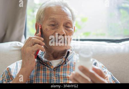 Healthy senior Asian man reading prescription bottle and talking on his cell phone at home. Medicine, online pharmacy and medical information concept. Stock Photo