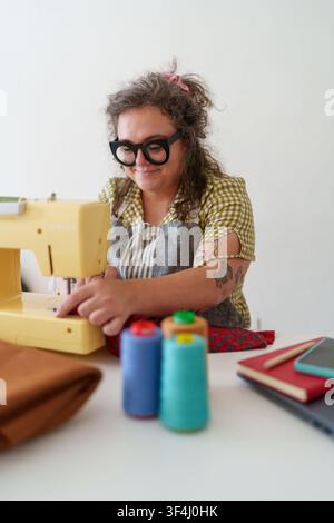 Woman focusing on her sewing machine, working on textile projects. Colorful thread spools and a notebook are on the table Stock Photo