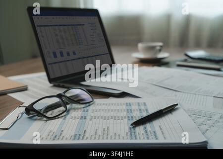 Laptop displaying financial report dashboard beside printed spreadsheets, pen, eyeglasses, coffee cup on desk, showing document review, data analysis, Stock Photo