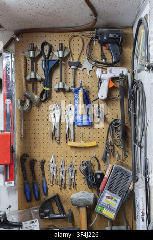 Various hand tools including vise grips, soldering and glue guns, sledge hammers hanging on light brown colored pegboard wall in home garage workshop Stock Photo