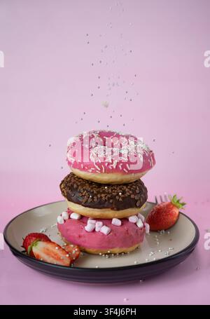 Stack of pink and chocolate glazed donuts decorated with sprinkles, served with fresh strawberries on a pastel pink background. Fun sweet dessert conc Stock Photo
