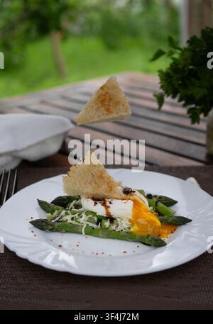Poached egg served with fresh green asparagus, grated cheese and toasted bread slices in midair on a white plate. Healthy gourmet breakfast outdoors. Stock Photo